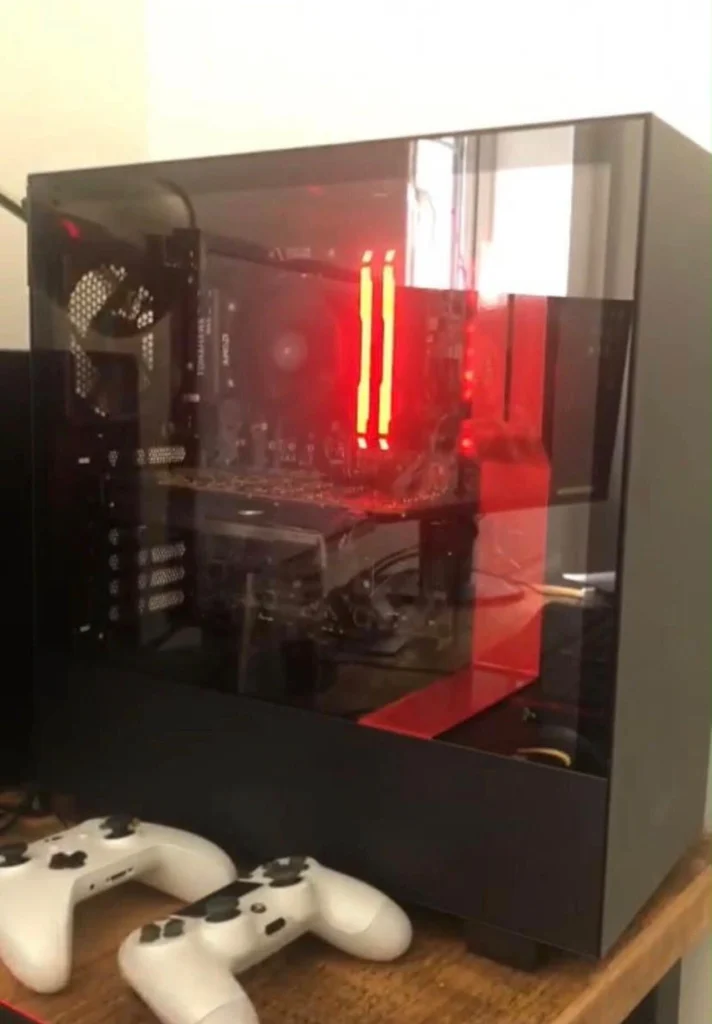 Black desktop gaming PC with a glass side panel, red internal lighting and two white game controllers placed in front.